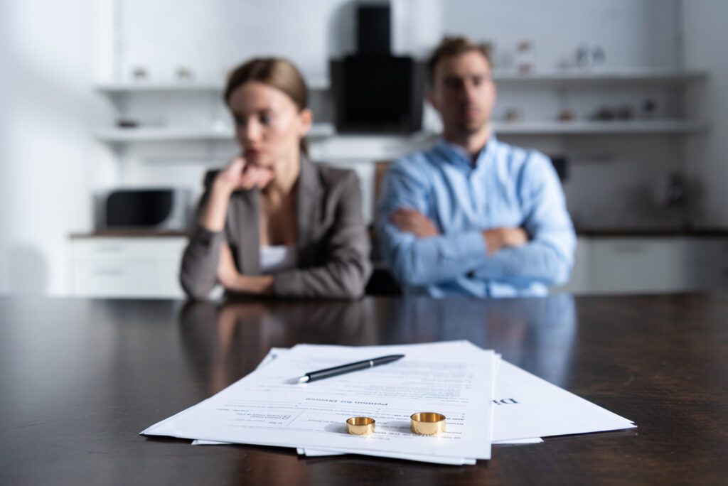 Cozakos and Centeno : A man and woman sit with crossed arms at a table, looking away from each other. In the foreground are legal documents, a pen, and two wedding rings.